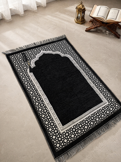 Black Ayahly prayer mat with woven arch design laid out beside a lantern and Quran stand